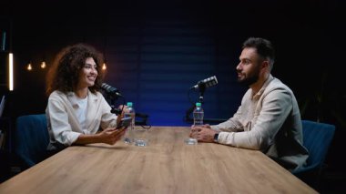 Kıvırcık saçlı genç bir bayan, modern bir kayıt stüdyosunda erkek bir konukla röportaj yapıyor. Her iki podcaster da canlı yayın sırasında profesyonel mikrofonlara konuşuyor.