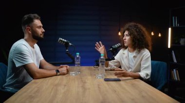 Kadın ve erkek, karanlık, profesyonel ışıklandırılmış bir stüdyoda mikrofonlu bir masada otururken canlı bir konuşma ve tartışma içerisindeki ses yayını veya radyo programını kaydediyor.