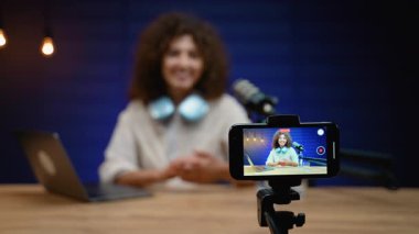 Genç kadın nüfuzlu kişi bir masada oturuyor ve bir podcast kaydederken veya cep telefonuyla canlı yayın videosu çekerken izleyicileriyle konuşuyor.
