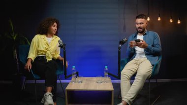 Genç bir adam ve kadın modern bir stüdyoda oturuyor ve birlikte bir ses yayını kaydediyor, mikrofonlara konuşarak dinleyicileri için canlı ve ilginç bir tartışma yapıyorlar.