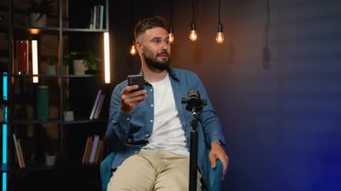Modern bir stüdyoda oturan, elinde akıllı bir telefonla profesyonel bir mikrofona konuşan, podcast yapan ya da sosyal medya izleyicileri için canlı yayın yapan genç bir adam.