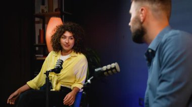 Kıvırcık saçlı, karizmatik bir bayan konuk canlı yayın programı için canlı bir röportaj sırasında canlı bir erkek sunucuyla konuşuyor. İkisi de modern bir kayıt stüdyosunda profesyonel mikrofonlara konuşuyor.