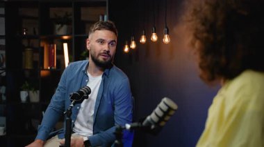 Karizmatik erkek radyo sunucusu canlı yayın sırasında bir kadın sunucuyla dostça sohbet ederken profesyonel bir mikrofona konuşuyor.