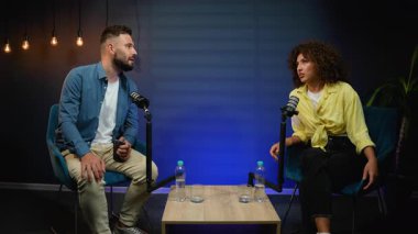 Erkek ve kadın canlı bir podcast programı sunuyor, profesyonel mikrofonlarla dolu bir masada otururken ve arka planda mavi ortam ışığı olan modern bir stüdyoda ilgi çekici bir konuşma yapıyorlar.