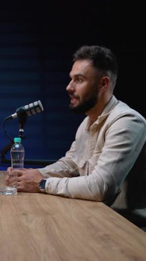 Stüdyo mikrofonuna konuşan yakışıklı sakallı bir adam podcast ya da radyo programı yaparken profesyonel bir kayıt stüdyosundaki karanlık bir arka planda, tahta bir masada oturuyor.