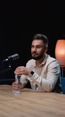 Genç sakallı bir adam karanlık bir stüdyoda ahşap bir masada oturmuş podcast röportajı sırasında profesyonel bir mikrofona konuşurken plastik şişeden bir bardak su dolduruyor.