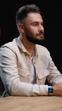 Tahta bir masada oturan ve röportaj sırasında mikrofona konuşan, elleri ve karanlık bir stüdyoda bir bardaktan su içen kendinden emin sakallı bir adam.