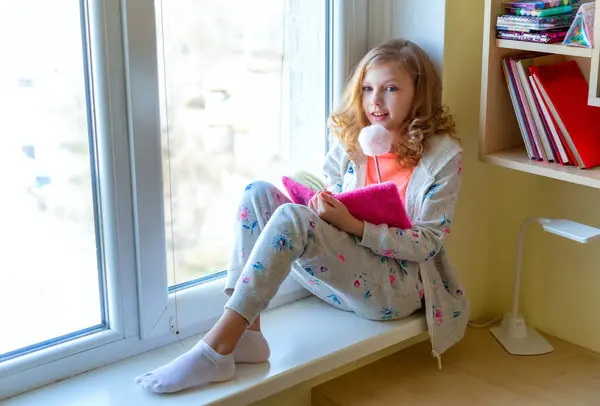 Beautiful little girl smiling and watching out the window. A child looks out the window.beautiful little teen girl sitting on the windowsill and writing something in a notebook with happy emotions