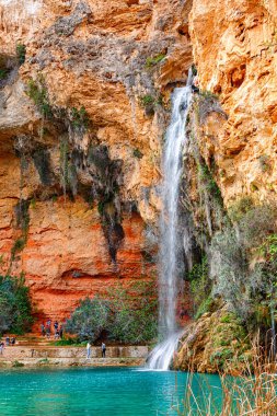 Ziyaretçiler kayalıkların arasına kurulmuş turkuaz bir havuza akan şelalenin çarpıcı manzarasının keyfini çıkarıyorlar..
