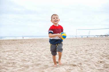 Neşeli genç bir çocuk kumsalda çömelmiş, bulutlu bir plaj gününde canlı bir plaj topuna uzanıyor..
