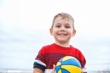 Neşeli genç bir çocuk kumsalda çömelmiş, bulutlu bir plaj gününde canlı bir plaj topuna uzanıyor..