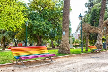 Spain, Valencia, March 29, 2024: A vibrant bench painted in rainbow colors sits in a lively park filled with lush trees and grass. clipart