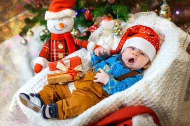 A sleepy baby wears a Santa hat and cozy outfit, nestled by holiday decorations and a cheerful snowman. clipart