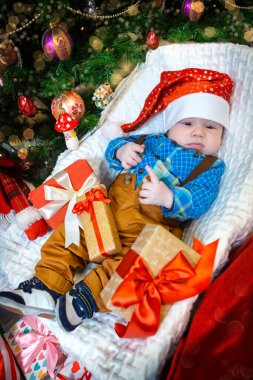 Şenlikli şapka ve mavi kareli gömlek giyen bir bebek, Noel ağacının renkli hediyelerinin arasında kutlamaya hazır bir şekilde oturur..
