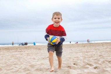 İspanya, Valencia, 29 Mart 2024: Neşeli genç bir çocuk kumsalda çömelmiş, bulutlu bir plaj gününde canlı bir plaj topuna uzanıyor..