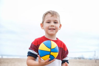 İspanya, Valencia, 29 Mart 2024: Neşeli genç bir çocuk kumsalda çömelmiş, bulutlu bir plaj gününde canlı bir plaj topuna uzanıyor..