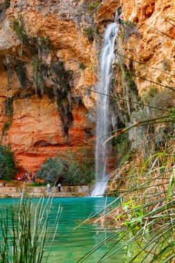 Ziyaretçiler kayalıkların arasına kurulmuş turkuaz bir havuza akan şelalenin çarpıcı manzarasının keyfini çıkarıyorlar..