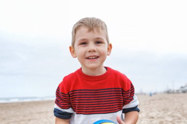 İspanya, Valencia, 29 Mart 2024: Neşeli genç bir çocuk kumsalda çömelmiş, bulutlu bir plaj gününde canlı bir plaj topuna uzanıyor..