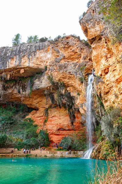 Resimli bir şelale, kayalık kayalıklar ve yemyeşil yeşilliklerle çevrili canlı bir turkuaz göle akar..