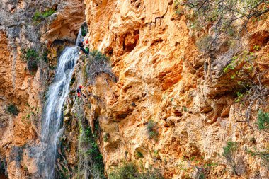 A daring climber ascends a rugged cliff adorned with patches of greenery while a waterfall cascades nearby.