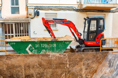 İspanya, Valencia, 29 Mart 2024: Kırmızı bir kazıcı aktif olarak yeşil bir çöp konteynırını işlek bir inşaat alanındaki kumla dolduruyor.