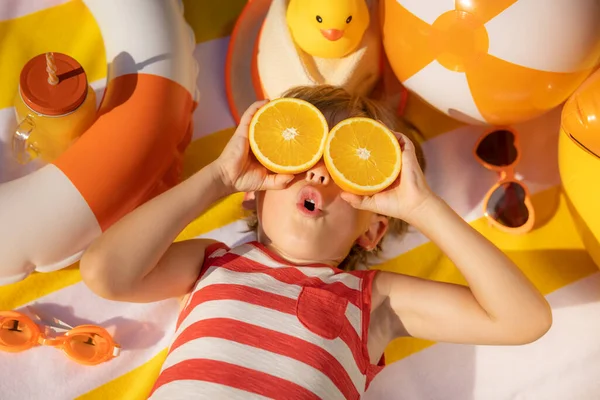 サングラスのようなオレンジの果実のスライスを保持する珍重された子供 子供の縞模様の黄色のTシャツビーチタオルの上に横たわるを着て 健康的な食事と夏休みのコンセプト — ストック写真
