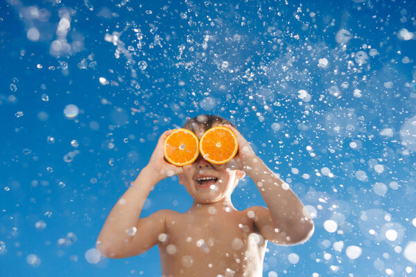 Happy child holding slices of orange fruit like sunglasses. Kid having fun against blue sky and splash background. Healthy eating and summer vacation concept