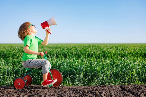 春の緑のフィールドで幸せな子供の自転車の屋外に乗る 青い夏の空を背景に大声で叫ぶ子供 — ストック写真