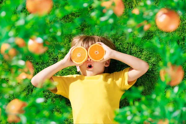 高傲的孩子手里拿着像太阳镜一样的橘子片 穿着黄色T恤的孩子躺在绿草上 健康饮食和暑假概念 — 图库照片