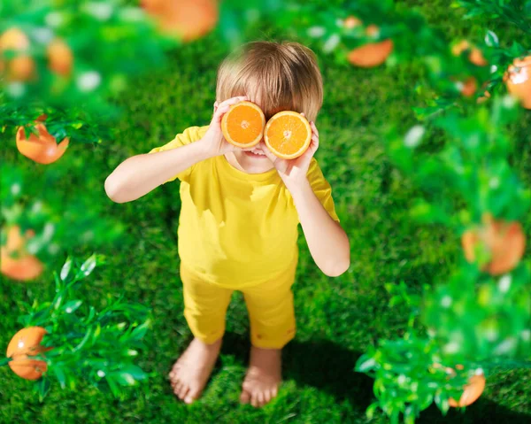 高傲的孩子手里拿着像太阳镜一样的橘子片 穿着黄色T恤的孩子站在绿草上 健康饮食和暑假概念 — 图库照片