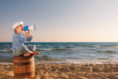 Çocuk denizci gibi davranıyor. Çocuk hoparlörü tutuyor. Çocuk sahilde eski fıçıda oturuyor. Yaz tatilindeki çocuk. Macera ve seyahat konsepti