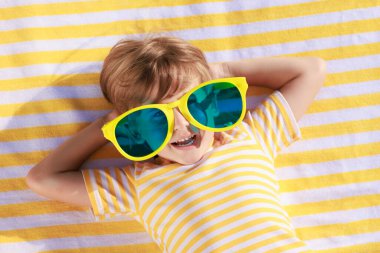 Mutlu çocuk plaj havlusunda yatıyor. Yaz tatilinde büyük güneş gözlüğü takan komik çocuk. Çocuk yaz tatilinde eğleniyor. Üst görünüm portresi