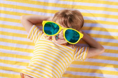 Mutlu çocuk plaj havlusunda yatıyor. Yaz tatilinde büyük güneş gözlüğü takan komik çocuk. Çocuk yaz tatilinde eğleniyor. Üst görünüm portresi