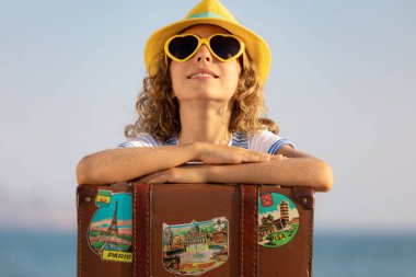 Eski bavullu mutlu çocuk gezgini. Küçük kız mavi denize ve gökyüzüne karşı. Yaz tatili ve seyahat konsepti