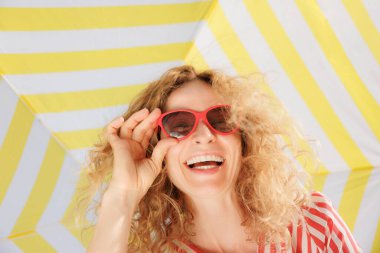 Sarı çizgili bir şemsiyenin altında mutlu bir genç kadın. Kameraya gülen bir kızın düşük açılı portresi. Güneş gözlüğü takan kadınlar yaz tatilinin tadını çıkarırlar.