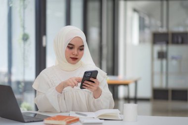 Beyaz tesettürlü mutlu ve güzel bir Müslüman iş kadını bir kafede dizüstü bilgisayarını kullanıyor.