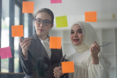 Şirket ekibi ofis alanında işbirliği yapıyor, cam duvara yapışkan notlar yapıştırarak yeni kavramları ve mevcut fikirleri canlandırıyor.