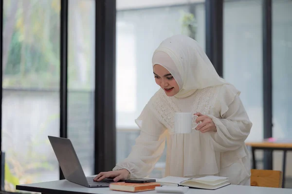 ラップトップを使用して現代的なオフィスでコーヒーを飲んでいる若いアジアのイスラム教徒のビジネス女性 — ストック写真
