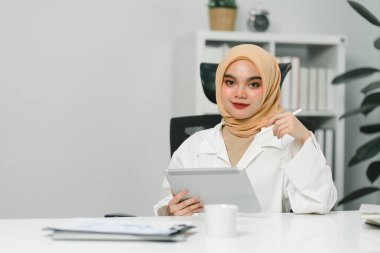 Güzel Asyalı Müslüman iş kadını modern ofisteki masada dijital tablet kullanıyor.