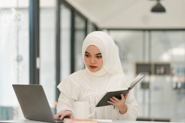 Güzel bir Müslüman iş kadını, modern bir ofiste masasında dijital bir laptop kullanıyor.