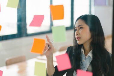 Bir iş kadını fikir alışverişinde bulunmak için post-it kullanır, beyin fırtınası kavramını gözler önüne serer. Yapışkan notlar cam bir duvara yerleştirilmiş.