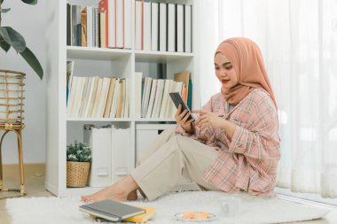 Güzel Müslüman kız tesettür takıyor, akıllı telefonuyla konuşuyor oturma odasında yerde oturuyor.