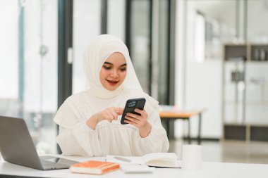 Müslüman iş kadını başörtüsü takıyor ve ofiste akıllı telefon kullanıyor.