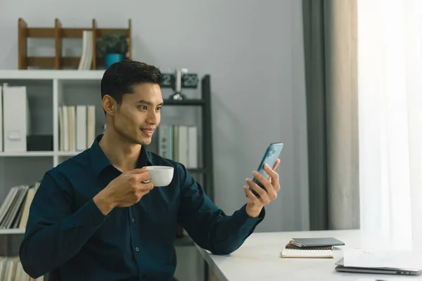 In the comfort of his home office, a young businessman can be seen multitasking, as he sips on a hot cup of coffee and deftly uses his smartphone