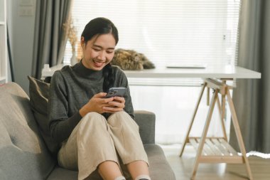 Çekici Asyalı kadın evdeki kanepede dinlenirken akıllı telefonunu kullanıyor.