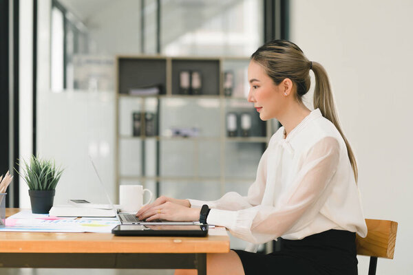 In a modern office, a beautiful Asian businesswoman is using a digital laptop at her desk