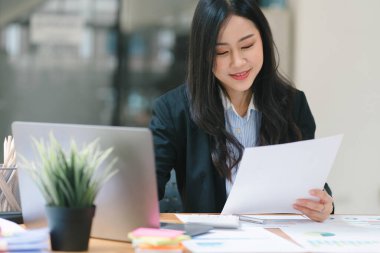 Asyalı güzel bir iş kadını, modern bir ofiste masasında dijital bir laptop kullanıyor.