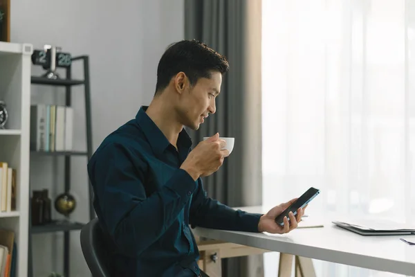 In the comfort of his home office, a young businessman can be seen multitasking, as he sips on a hot cup of coffee and deftly uses his smartphone