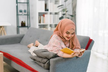 Evinin oturma odasındaki kanepede kitap okurken tesettüre giren gülümseyen Müslüman bir kadının portresi.