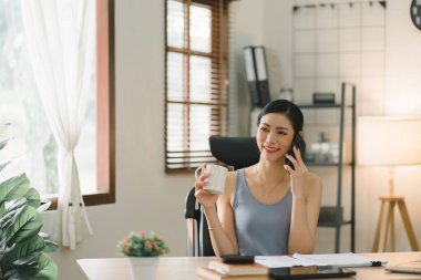 Görüntüde, neşeli genç bir iş kadını ev ofisinden çalışıyor, uzaktan çalışma konseptini örnek alıyor.